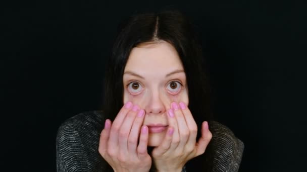 Young attractive brunette woman views her face in front of the camera as a mirror in black background. Room for text. — Stock Video