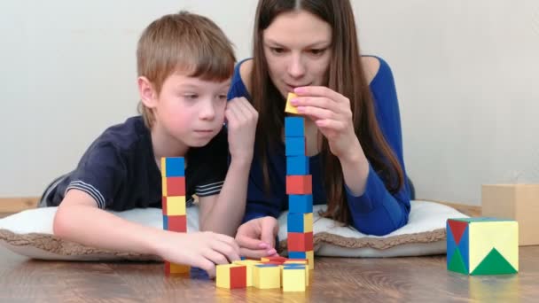 Bau eines Turms aus Blöcken. Mutter und Sohn spielen zusammen mit auf dem Boden liegenden farbigen Holzspielzeugklötzen. — Stockvideo