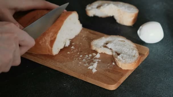 Κινηματογράφηση σε πρώτο πλάνο γυναικεία χέρια κόβοντας ένα κομμάτι ψωμί λευκό σε ξύλινη σανίδα σε μαύρο φόντο. Κάνοντας ένα σάντουιτς. — Αρχείο Βίντεο