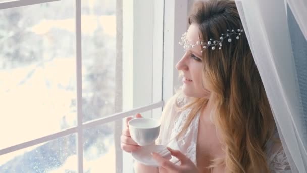 Joven chica rubia delgada sexy en lencería y tocador bebiendo un té y mirando en la ventana en invierno. Retrato de primer plano . — Vídeos de Stock