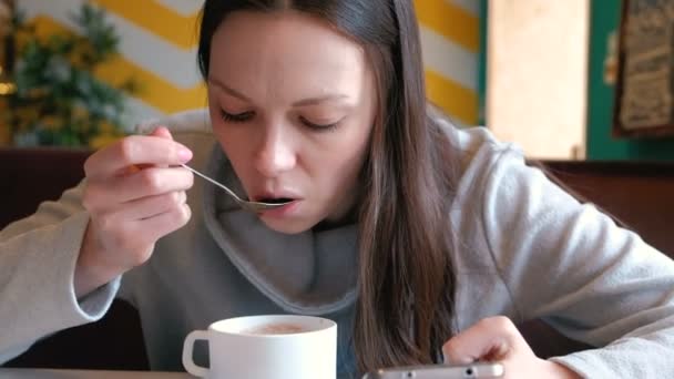 女性はカフェに座っている携帯電話の画面でスプーンとホット チョコレートを飲んでいます。. — ストック動画