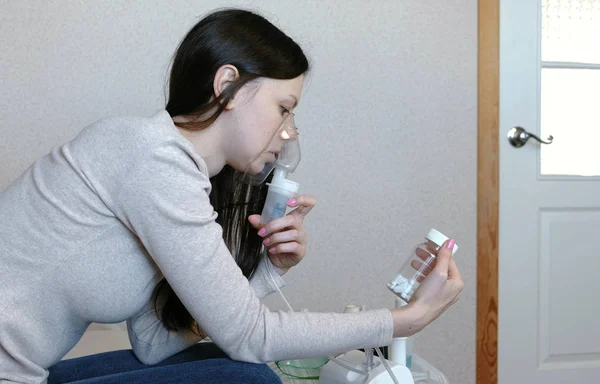 Use nebulizador e inalador para o tratamento. Jovem mulher inalando através de máscara inalador olha para um frasco de comprimidos. Vista lateral . — Fotografia de Stock