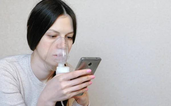 Use nebulizador e inalador para o tratamento. Mulher jovem inalando através da máscara do inalador e olhando para o telefone. Vista lateral close-up . — Fotografia de Stock