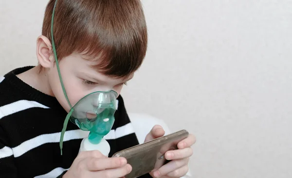 Use nebulizador e inalador para o tratamento. Closeup menino inalando através de máscara inalador e jogando o jogo em seu telefone móvel. Vista lateral . — Fotografia de Stock