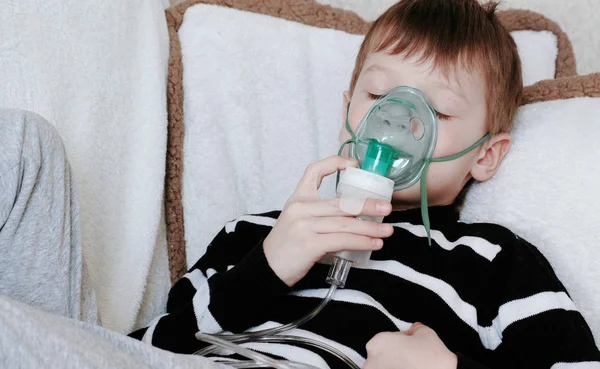 Przy pomocy rozpylacza i inhalator do leczenia. Chłopiec z cloused oczami wdechu przez inhalator maski leży na kanapie. — Zdjęcie stockowe