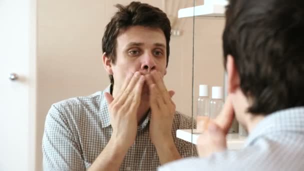 Mann cremt sich nach der Rasur ein, während er in den Spiegel schaut. brünett im karierten hellen Hemd. — Stockvideo