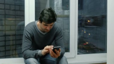 Genç adam bir pencere üzerinde oturan ve onun cep telefonu tarama.