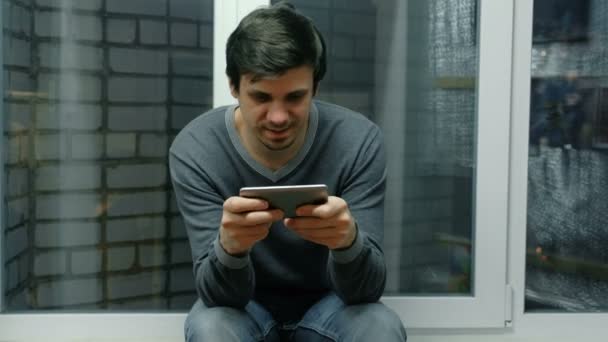 Ο άνθρωπος παίζει τα παιχνίδια σε ένα κινητό τηλέφωνο που κάθεται στο περβάζι. — Αρχείο Βίντεο