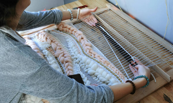 Weaving on a loom. Closeup womans hands running on a loom. Threading the shuttle through the strands of frame and fasten the yarn with scallop.