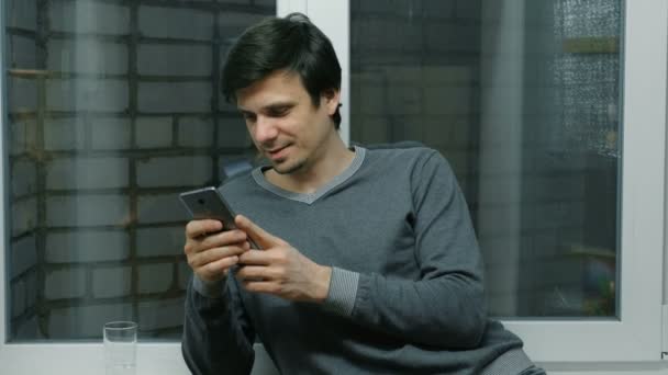 Homem está navegando páginas da internet no telefone celular em pé perto da janela na varanda . — Vídeo de Stock