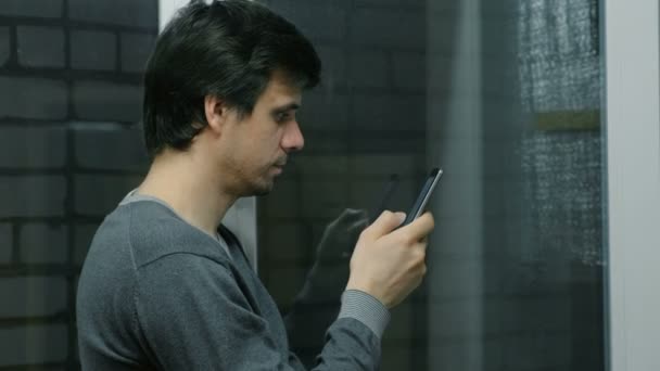 Homem está navegando páginas da internet no telefone móvel em pé perto da janela na varanda. Vista lateral . — Vídeo de Stock