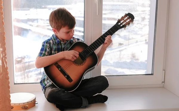 Играю на музыкальном инструменте. Недовольный мальчик играет на гитаре, сидя на подоконнике . — стоковое фото