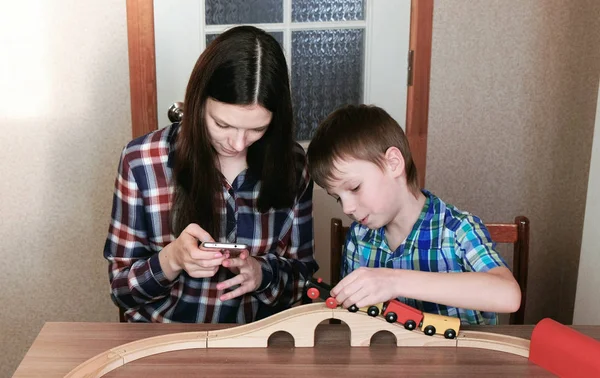 Együtt játszottak. Anya néz telefon és fia játszik egy fából készült vasúti vonat, a vagonok és a alagút ült az asztalnál. — Stock Fotó