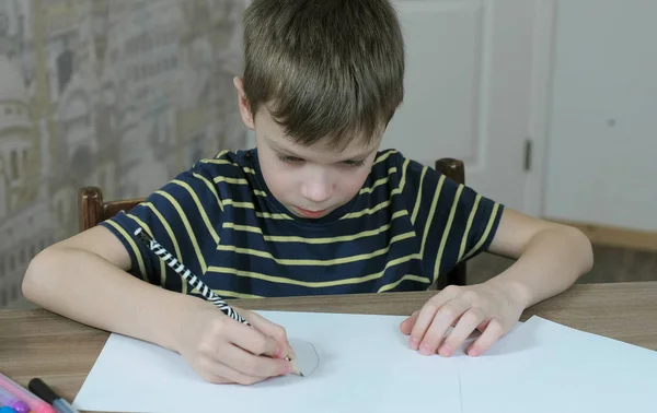 Boy of seven years in a striped t-shirt draws ovals with a simple pencil sitting at the table. — Stock Photo, Image