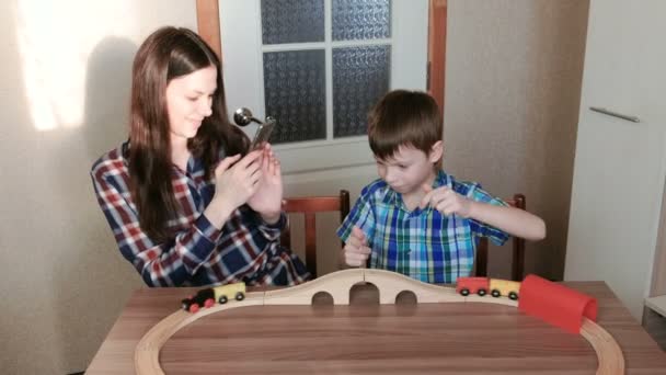 Brincando juntos. Mãe imagens uma foto no telefone como filho está jogando uma ferrovia de madeira com trem, vagões e túnel sentado à mesa . — Vídeo de Stock