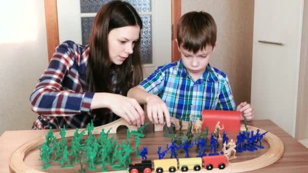 Zusammen zu spielen. Mutter und Sohn spielen eine hölzerne Eisenbahn mit Zug, Waggons und Tunnel mit Plastiksoldaten am Tisch. — Stockvideo