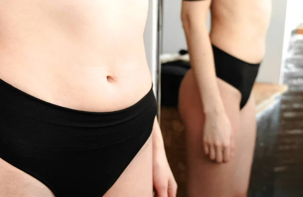 Thin young woman examines the folds on her stomach standing in front of a mirror in her underwear. Belly and arms close up. — Stock Photo, Image