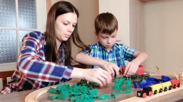 Jugando juntos. Mamá e hijo están jugando un ferrocarril de madera con tren, vagones y túnel con soldados de plástico sentados a la mesa . — Vídeos de Stock