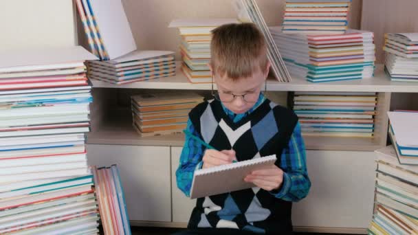 Семирічний хлопчик в окулярах малює щось в ескізі, що сидить серед книг . — стокове відео