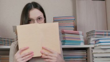 Gözlüklü genç esmer kadın kitap yüzünden gözlerini yükselterek bir kitap okur. Kitaplar arasında oturan.