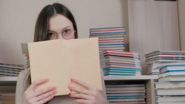 Giovane donna bruna in occhiali legge un libro, alzando gli occhi a causa del libro. Seduto tra i libri . — Video Stock