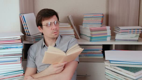 Pemuda berambut coklat berkacamata membaca sebuah buku dan berpikir duduk di antara buku-buku . — Stok Video