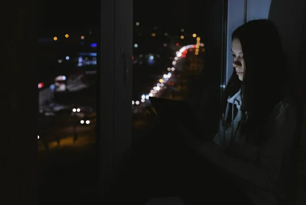 Женщина просматривает паутину на планшетном компьютере, сидя на подоконнике в темную ночь. Дорожный фон . — стоковое фото