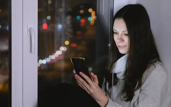 Frau surft mit Handy im Internet und sitzt in der dunklen Nacht auf der Fensterbank. Hintergrund Straße. — Stockfoto