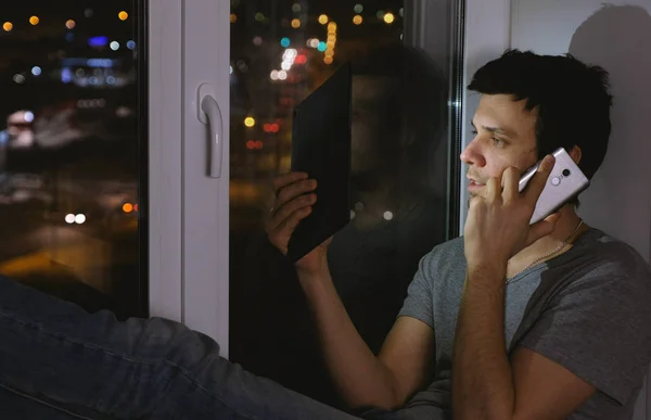 El hombre feliz está espiando el teléfono móvil y viendo la tableta. Sentado en el alféizar de la ventana en la noche oscura . — Foto de Stock