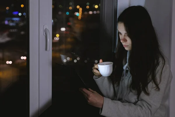 Mujer leyendo algo en la tableta y bebiendo té. Siéntate en el alféizar de la ventana en la oscuridad por la noche . — Foto de Stock