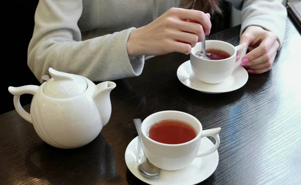 Close-up dames handen geroerd met een lepel suiker in thee. Theepot en kopjes op tafel. — Stockfoto