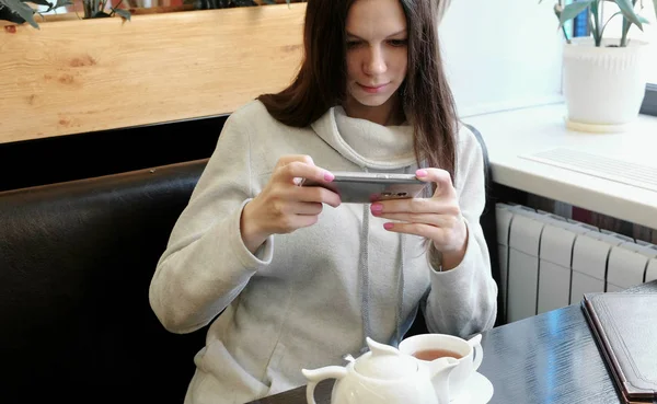 Девушка брюнетка фотографии чайник и Кубок в кафе на мобильный телефон. Вид спереди . — стоковое фото