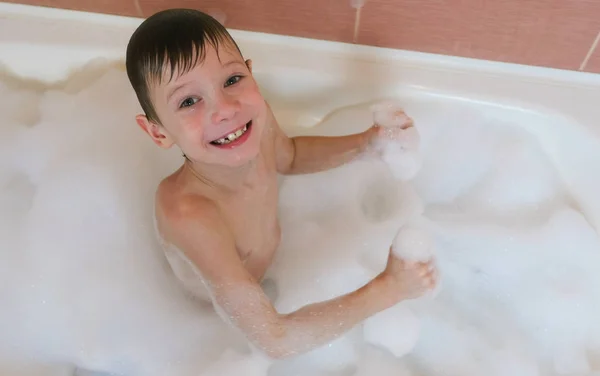 Boy 6-7 years sitting in the bath with foam looking into the camera and smiles. View top. — Stock Photo, Image