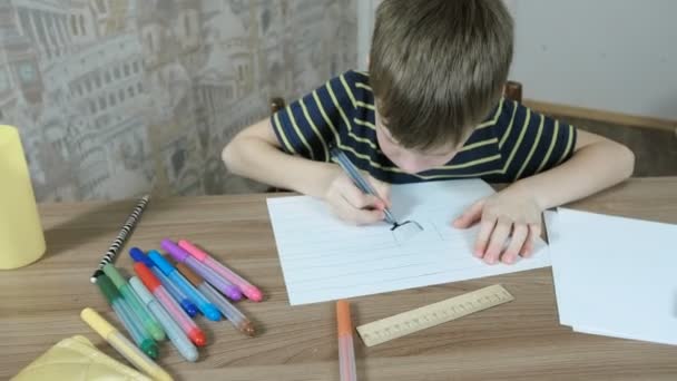 Garçon de 7 ans peint sa peinture avec un stylo feutre bleu en papier rayé assis près de la table . — Video