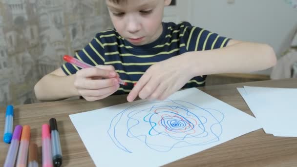 Jongen trekt spiraal van groene en oranje viltstift op wit papier zitten aan de tafel in zijn kamer. — Stockvideo