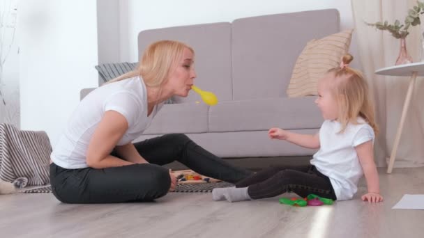 Atraente jovem loira mãe inflar um balão para sua filha encantadora sentado perto do sofá. Passatempo familiar . — Vídeo de Stock