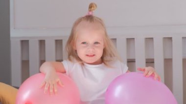 Yatak odasında pembe balonlu oturan ve gülümseyen küçük sevimli sarışın kız. Portre.