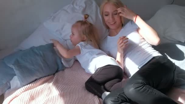 Schöne blonde Mutter und ihre kleine charmante Tochter liegen auf dem Bett und kuscheln, umarmen sich. — Stockvideo