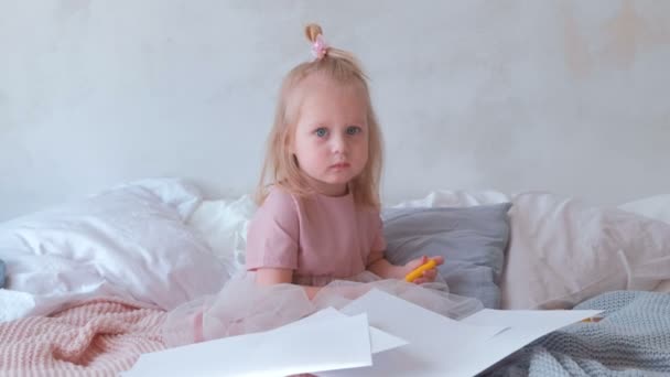 Little charming blond girl in pink dress holding a felt-pens and sitting on bed among paper. — Stock Video