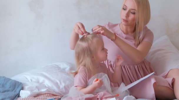 Giovane donna bionda attraente intreccia i capelli alla sua piccola figlia affascinante in abiti rosa mentre dipinge con feltro . — Video Stock