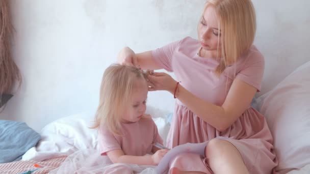 Joven y atractiva mamá rubia ata el cabello en trenzas a su pequeña hija encantadora con vestidos rosas mientras pinta con rotulador . — Vídeos de Stock