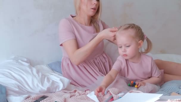 Молодая привлекательная блондинка мама привязывает волосы к косичкам своей маленькой очаровательной дочери в розовых платьях, пока она играет с войлочной ручкой — стоковое видео