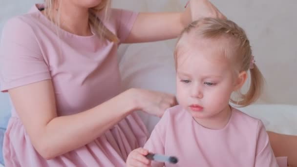 Mãe amarrar o cabelo em tranças para sua pequena filha encantadora em vestidos rosa, enquanto ela pintando com caneta de feltro . — Vídeo de Stock