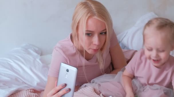 Joven mamá rubia atractiva tomar una foto divertida en el teléfono móvil con su pequeña hija encantadora en vestidos de color rosa. Chica escondida de la cámara . — Vídeo de stock