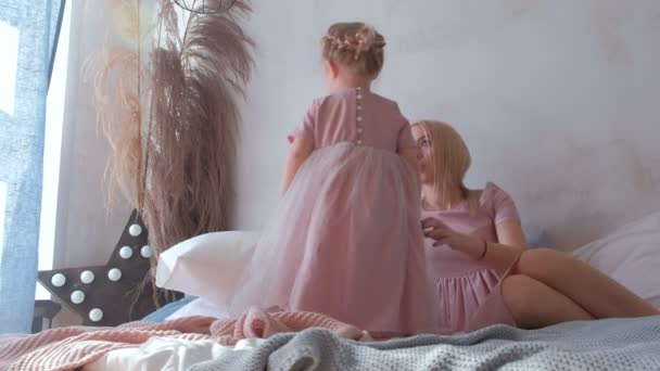 Joven y atractiva mamá rubia juega con su pequeña hija encantadora en vestidos rosados acostados en la cama . — Vídeo de stock