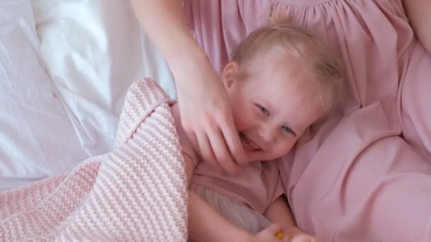 Piccola affascinante ragazza bionda gioca a nascondino con la mamma in abito rosa sdraiata sul letto . — Video Stock