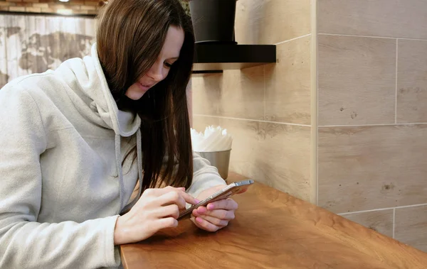 Улыбающаяся юная брюнетка в яркой рубашке, смотрящая в свой сотовый телефон, сидя в кафе . — стоковое фото
