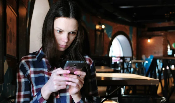 Αναστατωμένος γυναίκα στέλνει ένα μήνυμα ή να χρησιμοποιήσετε το internet στο τηλέφωνο κάθεται σε μια καφετέρια. Ντυμένος με ένα καρό πουκάμισο. — Φωτογραφία Αρχείου