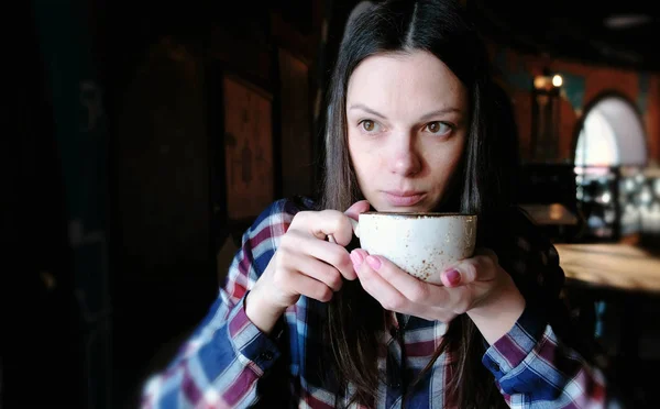 Mulher morena em camisa quadriculada bebendo chá de uma xícara grande. Mulher cansada tem um chá . — Fotografia de Stock