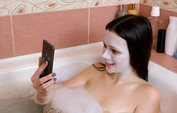 Brunette vrouw met een masker op haar gezicht ligt in badkamer en chattin door de mobiele telefoon en lachen. — Stockfoto